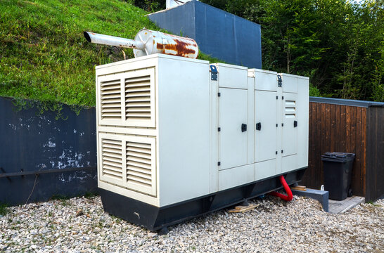 Large beige industrial soundproof generator with a rusted exhaust pipe mounted on top, positioned outdoors on a gravel ground