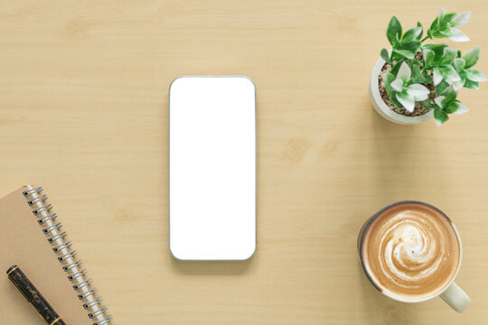 Top view flat lay of smartphone mockup with blank white screen on wooden desk, cup of coffee, notebook, pen, and green plant. Perfect workspace concept for business, modern office and design.