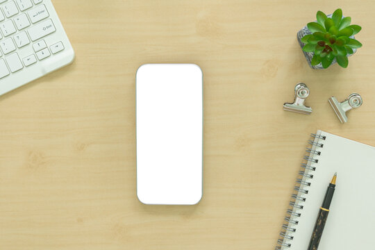 Top view of wooden desk with blank smartphone screen mockup, keyboard, plant, clips, and notebook with pen. Minimalist workspace template for business, tech, and digital marketing app display.