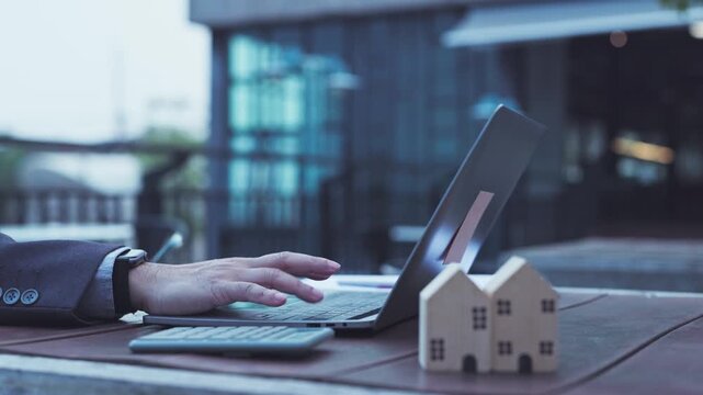 Businessman typing on laptop keyboard beside wooden miniature house models calculator on outdoor table calculate property mortgage loan rates, investment cost, budget plan, real estate insurance.