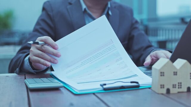 Real estate agent analyzing mortgage loan document paperwork near laptop computer, calculator, wooden house model on desk outdoors.