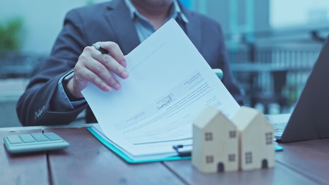 Real estate agent reviewing mortgage contract documents beside house model miniatures, calculator, laptop outdoors.