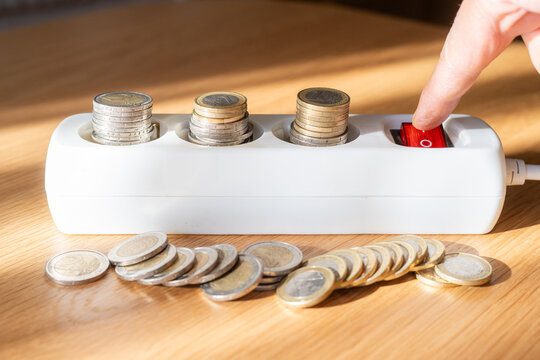 Finger pressing the red switch of an electrical power strip with stacked euro coins representing high costs, illustrating energy efficiency and potential savings on bills