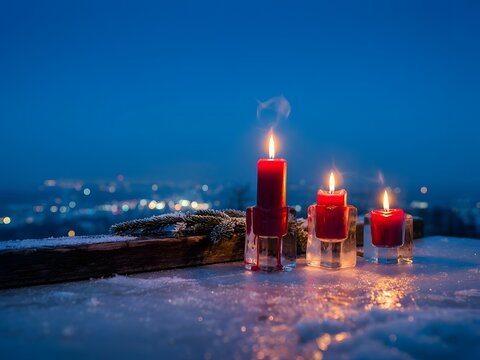 Three glowing red advent candles with smoke rising on a frosty winter evening with bokeh city lights in the background