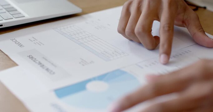 Adult right hand is entering frame, tracing invoice on desk and tapping laptop to verify totals