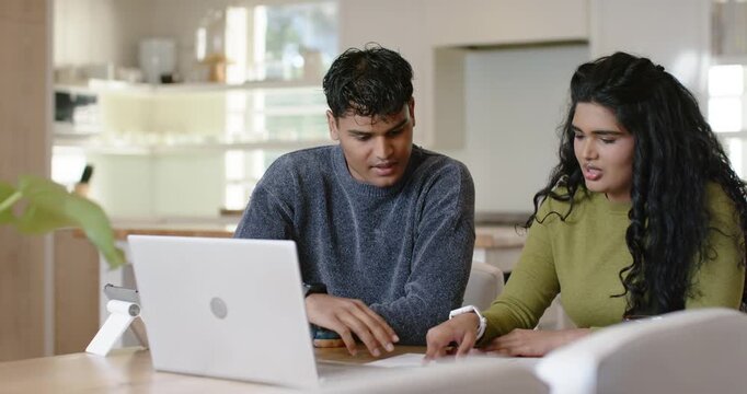 Diverse couple spotting open laptop, pointing and comparing papers at kitchen table, making plan