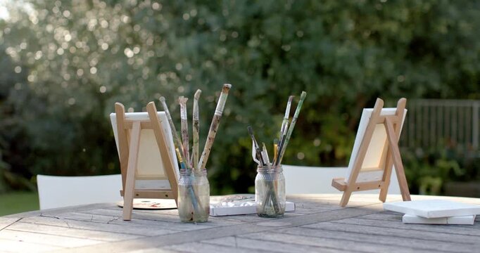 Camera is widening, shifting focus, revealing easels with brush jars on garden table to frame scene