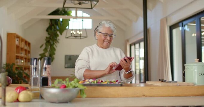 Senior woman at kitchen island prepping vegetables on tray while checking recipe alert on red phone