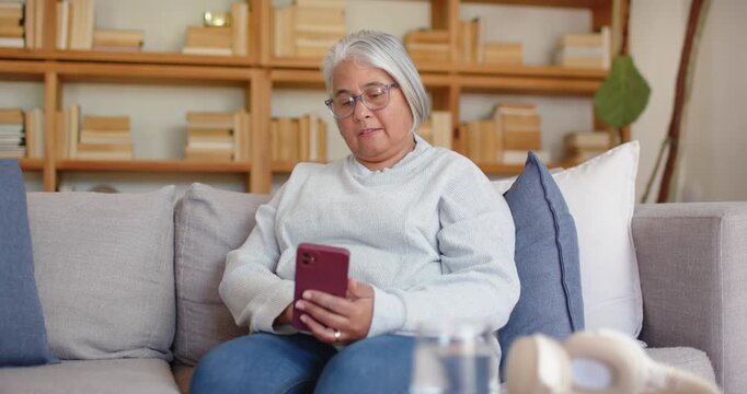 Senior female seeing notification, tapping and scrolling smartphone on couch, headphones near