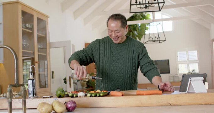 Asian man in green sweater consulting tablet recipe, pouring oil onto vegetables on wooden island