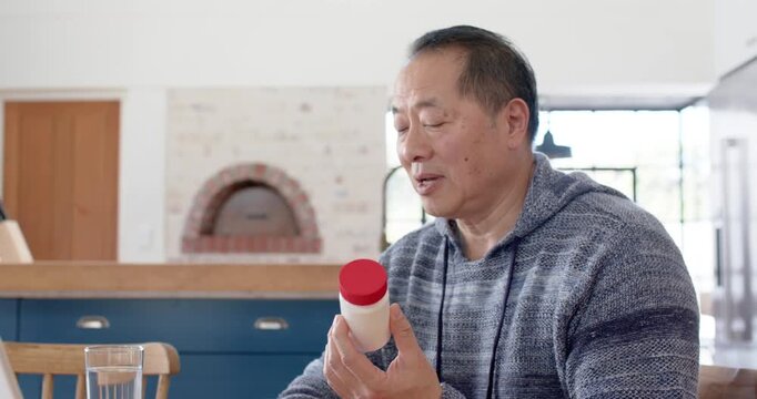 Asian man 50s in striped hoodie holding white bottle verifying label from laptop at kitchen table