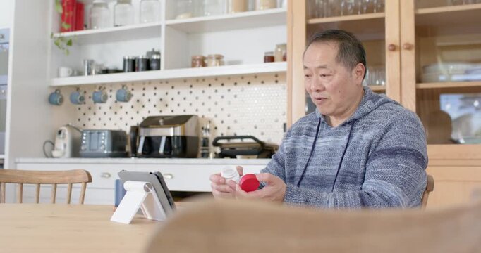 Senior Asian man at kitchen table following tablet, opening white bottle with red lid, copy space