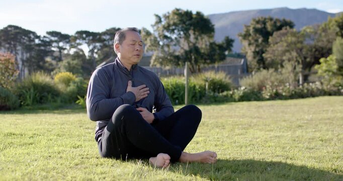 Asian senior man meditating on grass, placing hands on chest and abdomen, breathing slowly