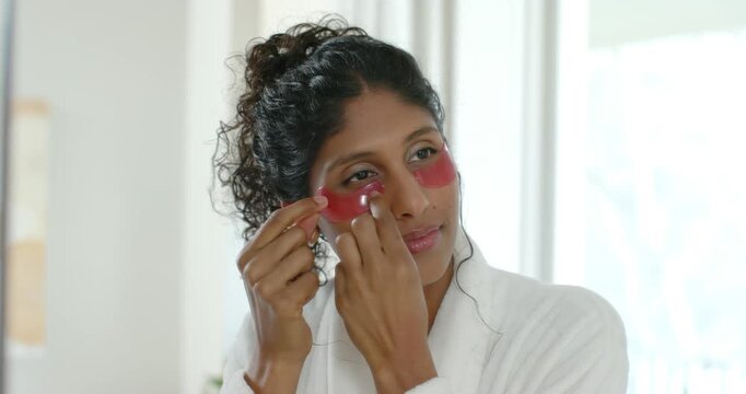 African American woman placing hydrogel patches, pressing under eyes at vanity for skincare in robe
