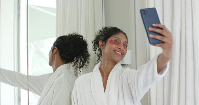 African American woman checking red gel patches at vanity, posing for selfies with phone in robe