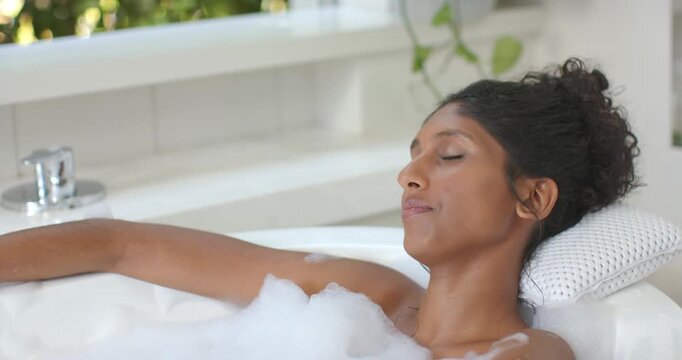 African American woman reclining in bubble bath foam on cushioned pillow, opening eyes to sunlight