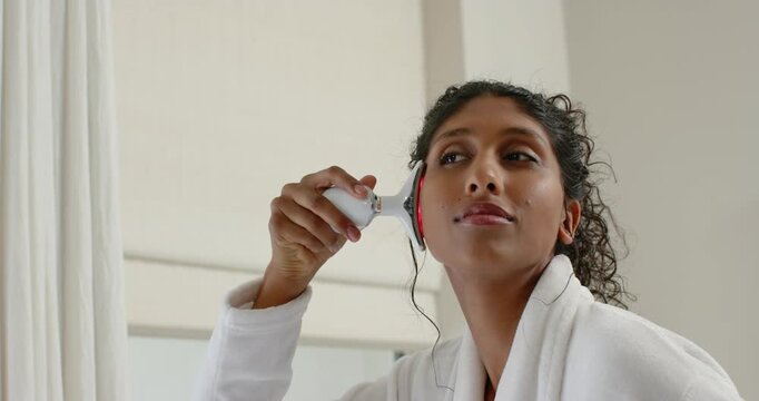 African American woman in robe pressing LED wand on right-cheek at vanity applying serum for skin