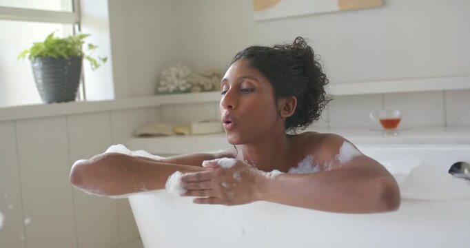 African American woman scooping foam, blowing bubbles, rubbing neck in white tub by window