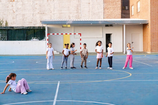 Group of diverse elementary school children standing together on a schoolyard while one child is sitting isolated on the ground