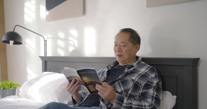 Senior Asian man opening paperback and reading, turning pages and smiling at story on bed