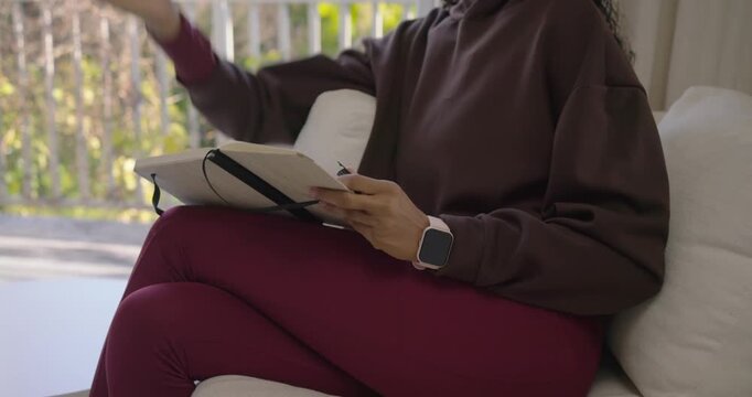 Adult Indian woman lifting notebook and writing plans with pen, smartwatch on white sofa near deck