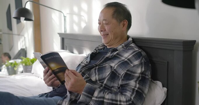Mature Asian man holding paperback open, sitting against dark headboard, turning pages for rest