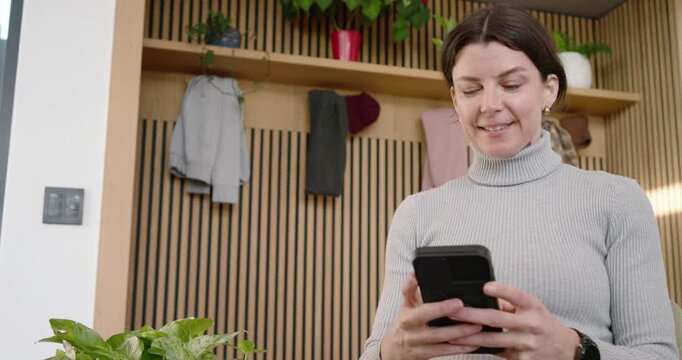 Woman in 30s wearing gray sweater reading message, tapping phone, smiling in front of wooden slats