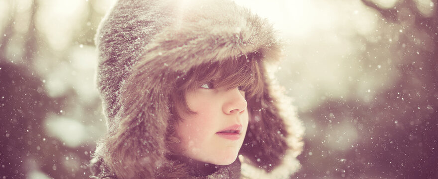 Young child looking away, wearing a furry trapper hat, experiencing a sunny winter snowfall outdoors