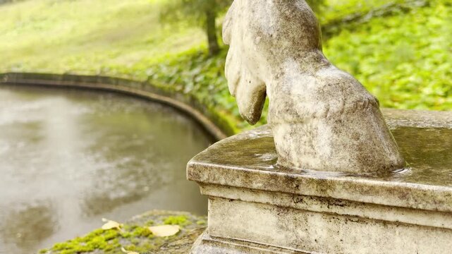 A wet stone centaur statue drips raindrops beside a quiet river. The peaceful autumn rain falls softly over green moss in Pavlovsk Park, Russia, captured in serene slow motion.