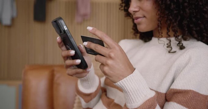 African American woman in cream sweater on couch bringing card to phone, tapping