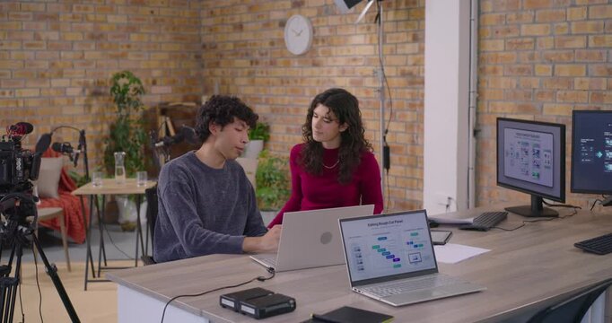 Studio colleagues reviewing man gesturing at laptop urging woman in red matching print with monitor