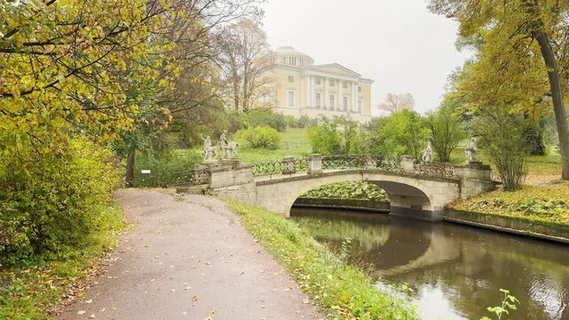 POV walkthrough glides along a quiet path toward Centaur Bridge and Pavlovsk Palace in slow motion. The autumn scenery inspires a peaceful and majestic mood. Shot in Pavlovsk Park, Russia.