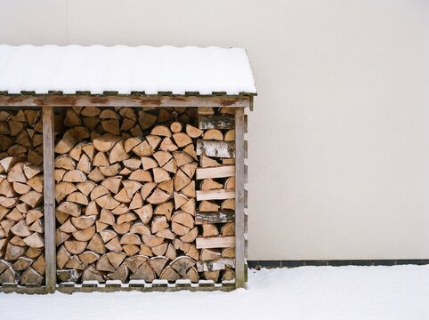 Wooden log store filled with split firewood under snow by a house wall