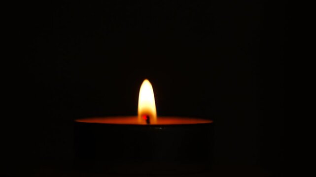 Lone memorial candle burning low in the darkness with an amber glow on the wax rim, quiet solemn composition for All Saints Day and All Souls Day, concept of remembrance, mourning and honoring loved 