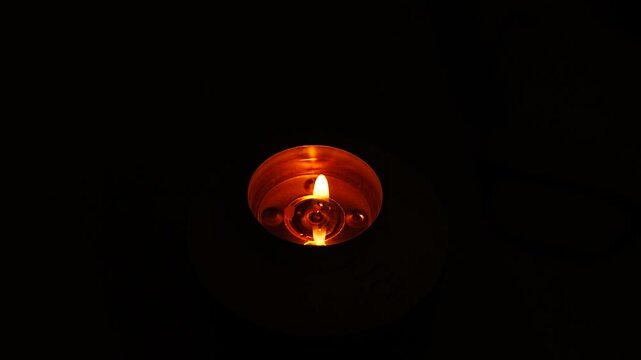 Single tea light burning steadily in a completely dark room, the only source of light glowing orange from above, concept of power outage, blackout, emergency light and energy crisis