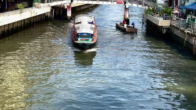 A passenger boat travels through a narrow urban canal, creating ripples and foamy waves on the water as it passes concrete embankments and riverside walkways in Bangkok, Thailand.