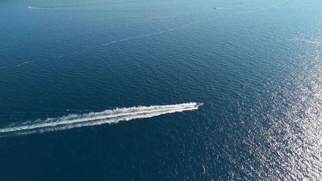Off the coast of Polignano a Mare, a motorboat races diagonally across sun-glittered Adriatic water, drawing a bright foamy trail. The high angle with open sea all around suits leisure, motion and su