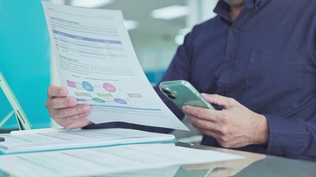 Focused corporate employee examines technical system architecture workflow document, cross-checking operational process charts beside desk tablet device.