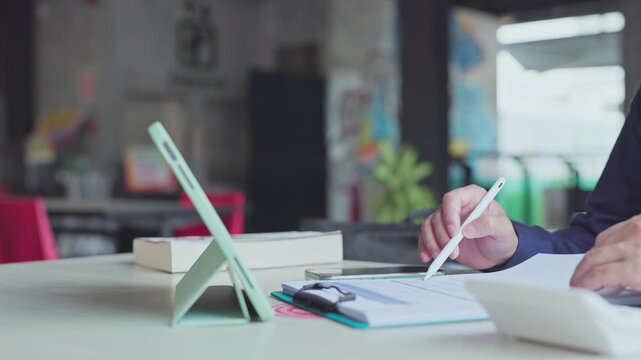 Professional holding digital pen over paper documents beside tablet computer, reviewing financial report, analyzing business data, calculating budget at office desk.