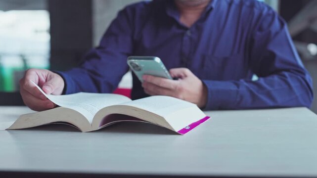 Mid adult man reading textbook open across desk checking mobile phone message indoors.