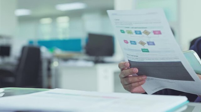 Software developer analyzing printed flowchart documentation source code sheets beside mobile phone in bright corporate office workspace.