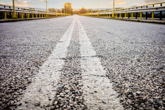 Empty asphalt country road with double white dividing lines leading towards the horizon under a bright morning light with blurred yellow safety barriers.