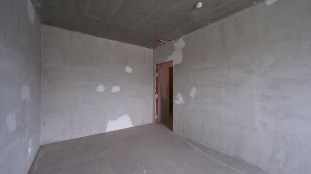 Sparse unfinished room with raw concrete surfaces. rough plaster walls, exposed ceiling, and distant doorway convey early construction stage