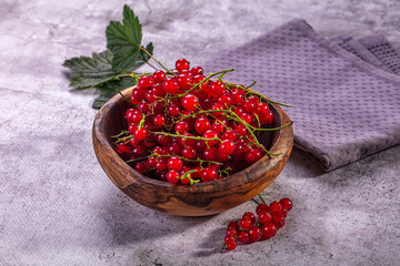 Red currant berries piled inside wooden bowl beside green leaf on textured grey countertop surface, fresh summer fruit harvest © Andrei Starostin