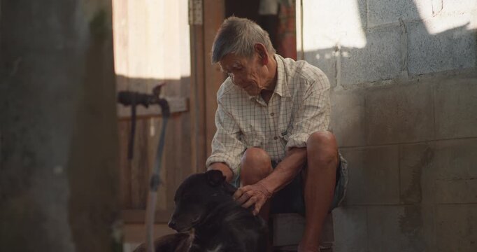 A man who is senior sits on his doorstep petting a black dog, barefoot in the warm evening sunlight of a weathered old house.