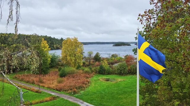 Close op view of Swedish flag waving beside autumn trees overlooking Baltic Sea on cloudy day. Sweden.