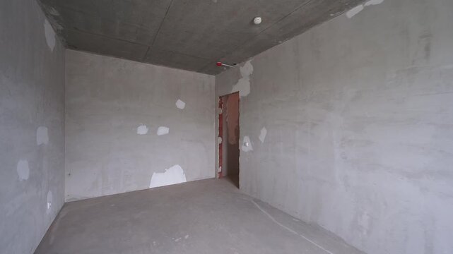 Sparse unfinished room with raw concrete surfaces. rough plaster walls, exposed ceiling, and distant doorway convey early construction stage