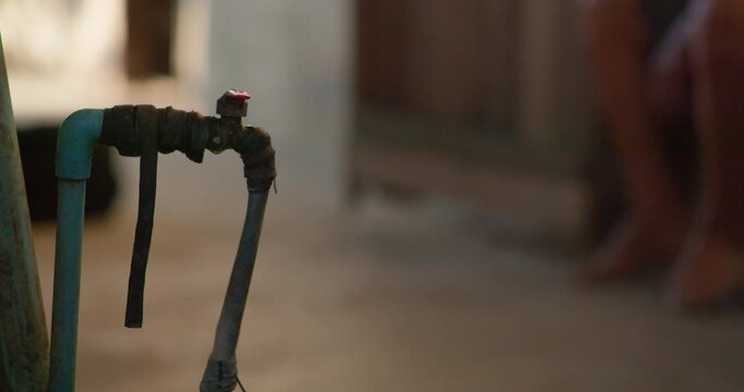 Water drips from a weathered, taped-up pipe tap in a dim rustic alleyway, worn pipes bound together with old rubber hose.