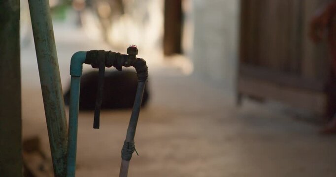 Water drips from a weathered, taped-up pipe tap in a dim rustic alleyway, worn pipes bound together with old rubber hose.
