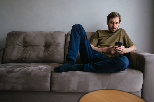 Man relaxing on a comfortable sofa, leaning back against the cushions and interacting with his mobile phone, browsing online content or sending messages in a casual setting